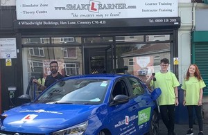 Driving instructor Tommy Sandhu and two students in bright green shirts standing next to a blue electric vehicle, one holding a charger for the car